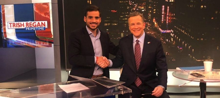 Armando Armas estrechando la mano de un hombre en un estudio de televisión con el logotipo de Trish Regan Primetime.