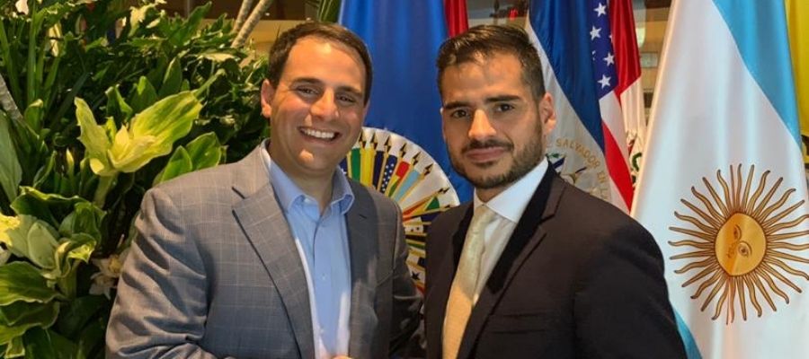 Armando Armas junto a un hombre vestido de traje frente a banderas de Venezuela, Estados Unidos y Argentina.