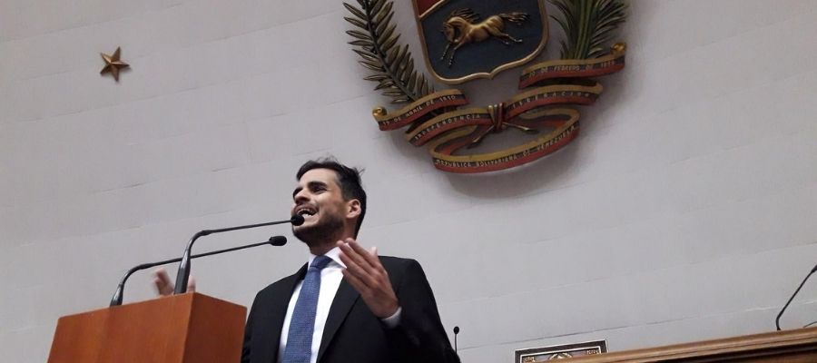 Armando Armas hablando desde una tribuna con micrófonos y el escudo de Venezuela al fondo.