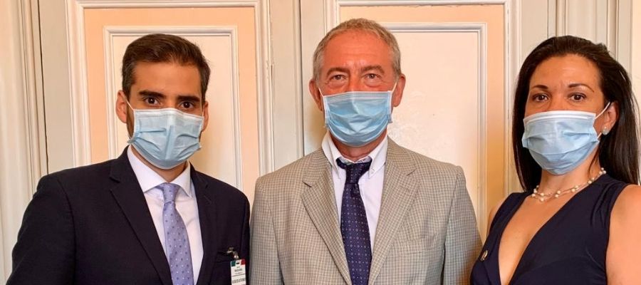 Armando Armas con traje oscuro y mascarilla junto a dos personas en una fotografía institucional.