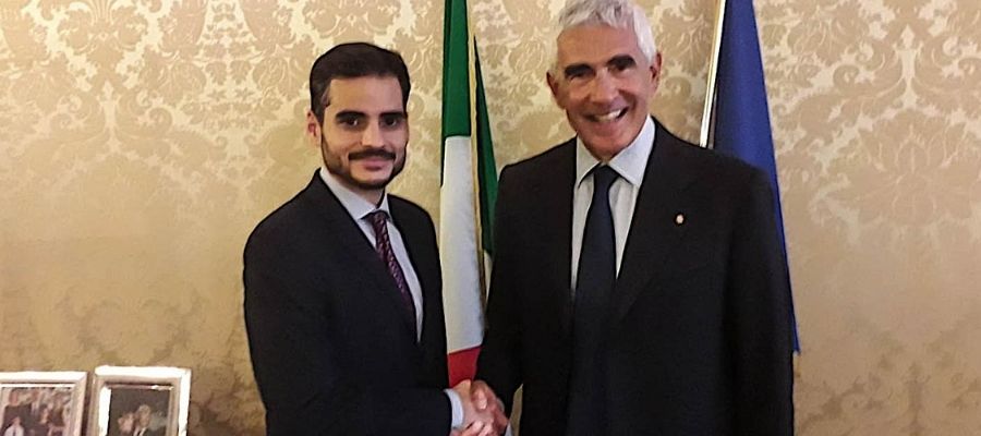 Armando Armas estrechando la mano de un hombre de traje frente a banderas de Italia y la Unión Europea.