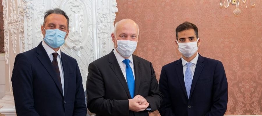 Armando Armas con traje azul y mascarilla junto a dos hombres durante un encuentro institucional.