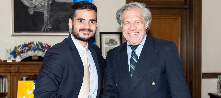 Armando Armas junto a un hombre de traje oscuro en una oficina con banderas internacionales.