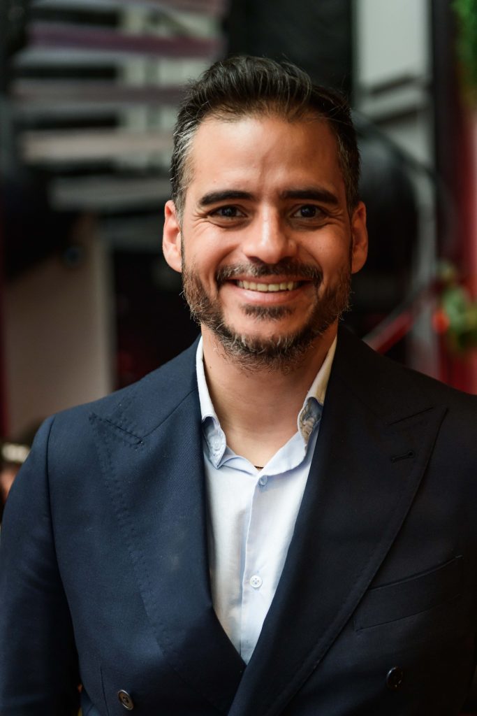 Retrato de Armando Armas sonriendo, vestido con traje azul oscuro y camisa clara.