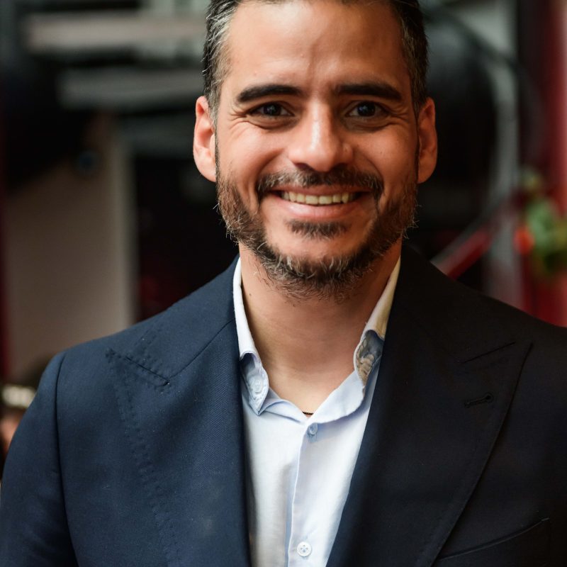 Retrato de Armando Armas sonriendo, vestido con traje azul oscuro y camisa clara.